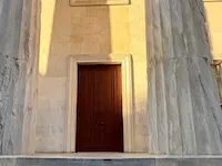 A dark red door centered in a close-up photo of a stone building with imposing pale beige columns